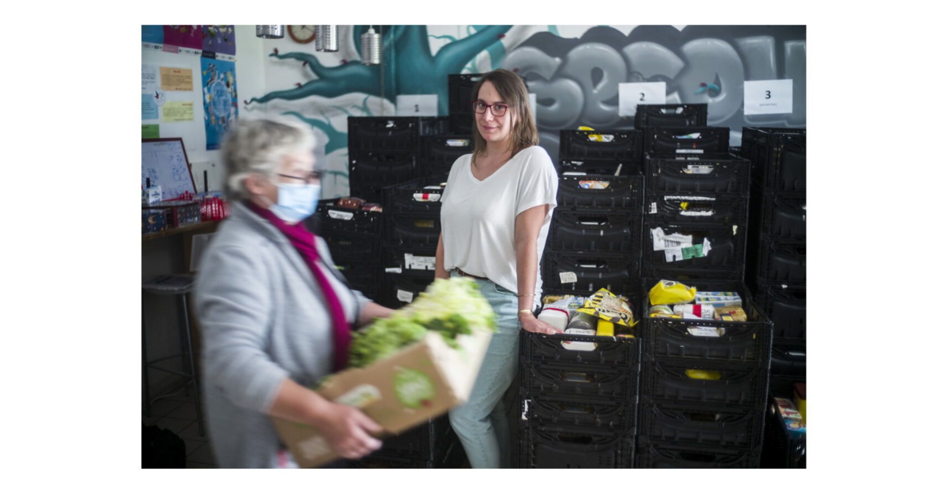 2 bénévolesdans un local de distribution de produits alimentaires, l'une posant debout, l'autre transportant une caisse de salade